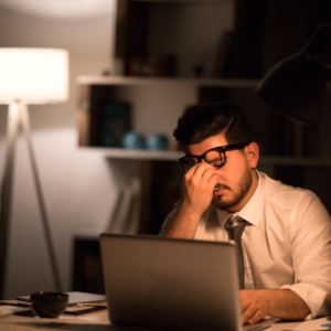 A bearded man sat at a laptop with his head in his hands, looking exhausted 