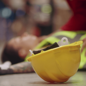 Man unconscious on floor wearing a high-vis jacket with a yellow helmet in the foreground 